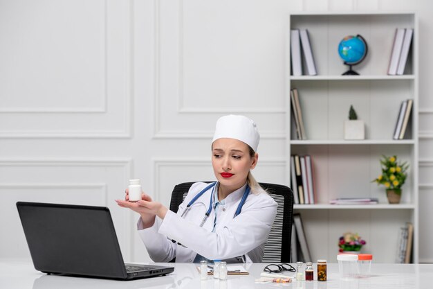 Docteur médical assez mignon en blouse blanche et chapeau avec ordinateur tenant des pilules dans les mains