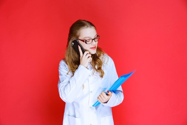 Docteur portant des lunettes et tenant un dossier bleu parlant au téléphone.