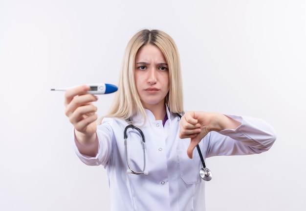 Docteur strict jeune fille blonde portant un stéthoscope et une robe médicale tenant un thermomètre à la caméra son pouce vers le bas sur fond blanc isolé