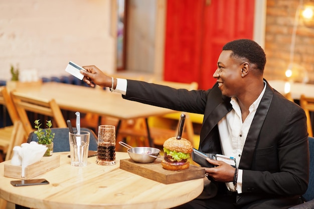 Photo gratuite donne la carte de crédit du portefeuille au serveur payant par la nourriture respectable jeune homme afro-américain en costume noir assis au restaurant avec un délicieux double burger et une boisson gazeuse