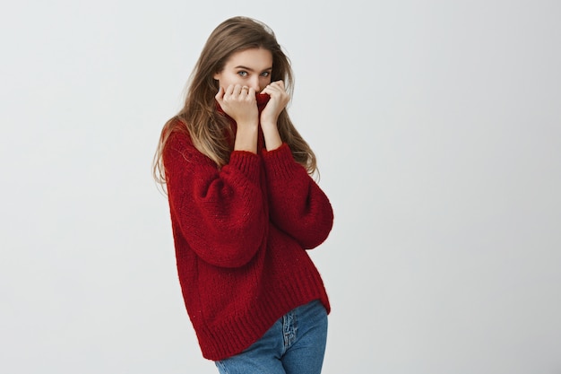 Donnez-moi des câlins pour me réchauffer. Portrait de jolie femme européenne mignonne se cachant le visage dans le col du pull en regardant en se tenant debout Une fille se fige à cause d'un radiateur cassé