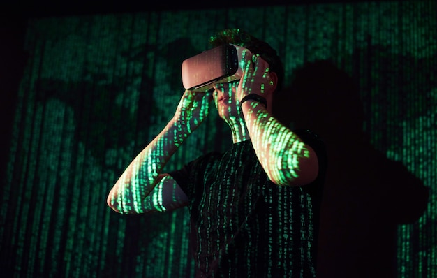 La double exposition d'un homme caucasien et d'un casque de réalité virtuelle VR est vraisemblablement un joueur ou un pirate informatique déchiffrant le code dans un réseau ou un serveur sécurisé, avec des lignes de code en vert