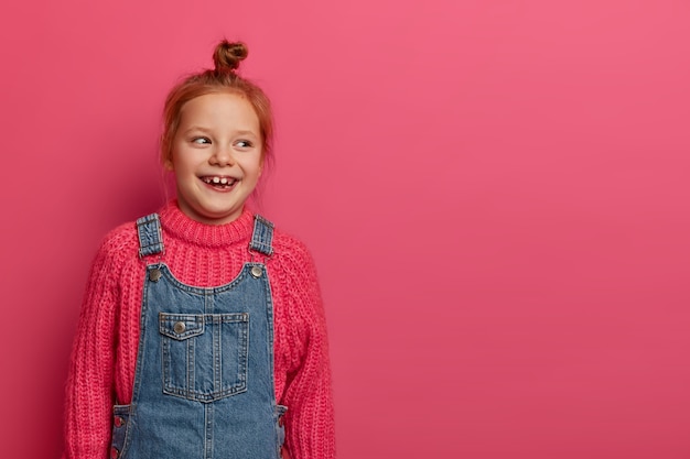 Douce jolie fille sourit positivement et regarde de côté, a un nœud de cheveux roux, un sourire à pleines dents, porte un pull et une salopette tricotés, aime les jours et les week-ends libres de l'école, rit de quelque chose de drôle.