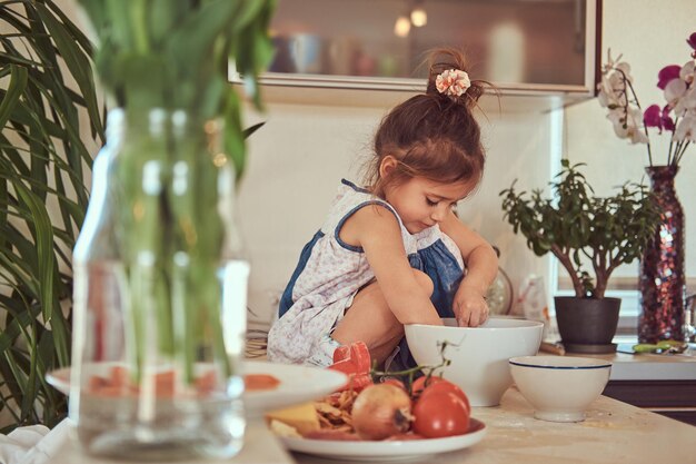 Douce petite fille mignonne apprend à cuisiner un repas dans la cuisine tout en étant assise sur un comptoir.