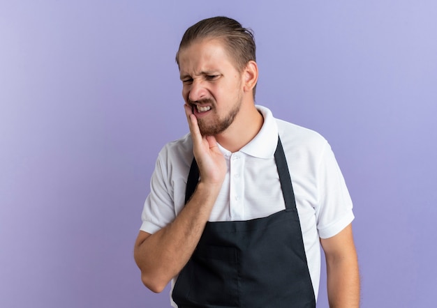 Douleur jeune beau coiffeur en uniforme mettant la main sur la joue souffrant de maux de dents isolé sur violet avec copie espace