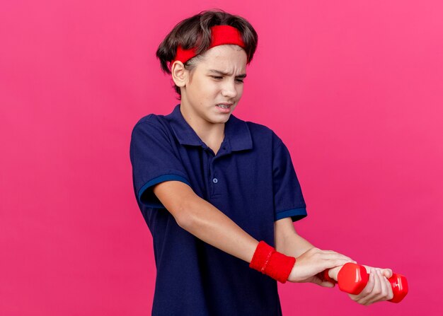 Douleur jeune beau garçon sportif portant un bandeau et des bracelets avec un appareil dentaire tenant un haltère en regardant et en mettant la main sur le poignet isolé sur un mur cramoisi avec espace de copie