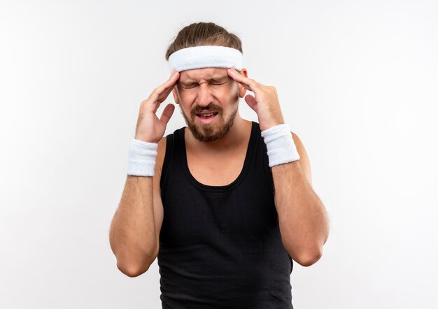 Douleur jeune bel homme sportif portant un bandeau et des bracelets mettant les mains sur les tempes souffrant de maux de tête avec les yeux fermés isolés sur un mur blanc