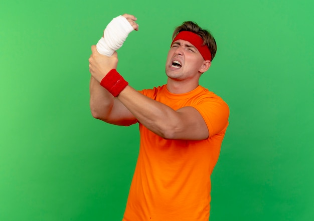 Douleur jeune bel homme sportif portant un bandeau et des bracelets soulevant et tenant son poignet blessé enveloppé de bandage isolé sur vert avec espace copie