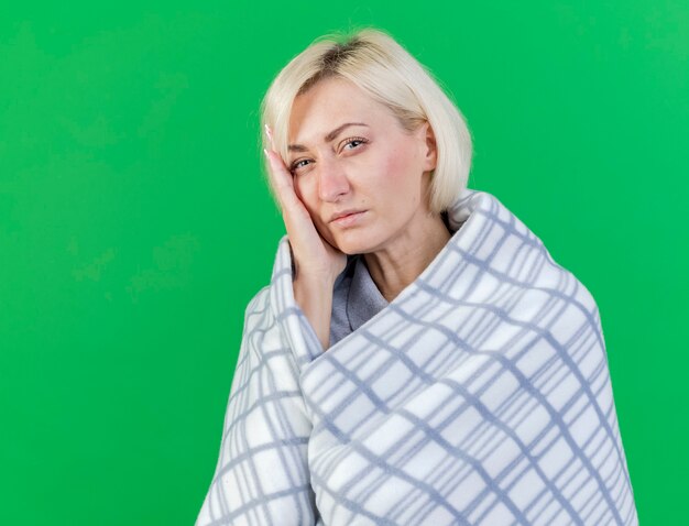 Douleur jeune femme malade blonde enveloppée dans un plaid met la main sur le visage isolé sur mur vert