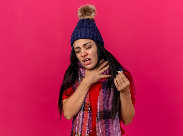 Douleur jeune femme malade portant chapeau d'hiver et écharpe tenant capsule et sa gorge avec les yeux fermés isolé sur mur rose