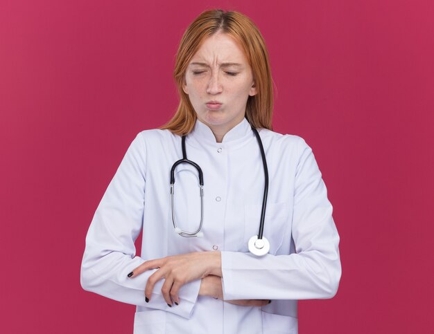 Douleur jeune femme médecin gingembre portant une robe médicale et un stéthoscope gardant les mains sur le ventre avec les yeux fermés isolés sur un mur cramoisi