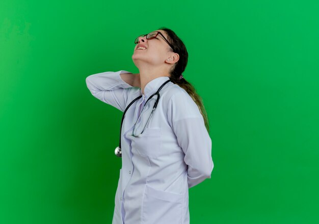 Douleur jeune femme médecin portant une robe médicale et un stéthoscope et des lunettes debout en vue de profil mettant les mains derrière le cou et derrière le dos isolé sur un mur vert avec espace copie