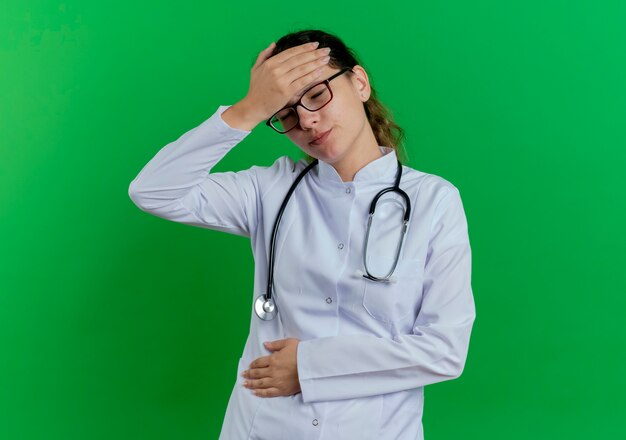 Photo gratuite douleur jeune femme médecin portant une robe médicale et un stéthoscope et des lunettes tenant la tête et le ventre avec les yeux fermés