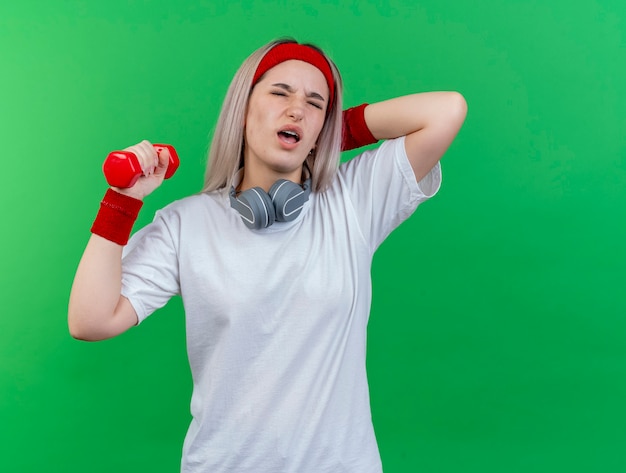 Douleur jeune femme sportive avec des accolades portant un bandeau et des bracelets avec des écouteurs sur le cou met la main sur le cou derrière et détient un haltère isolé sur un mur vert