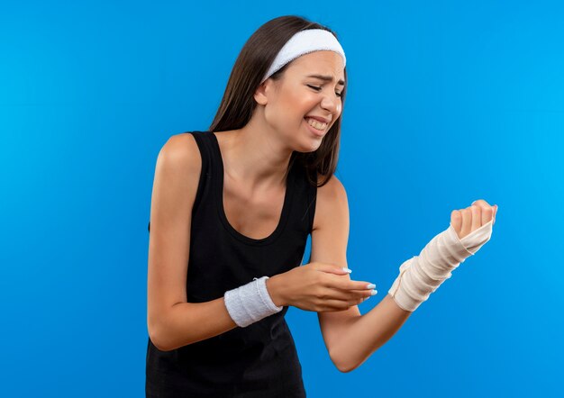 Douleur jeune fille assez sportive portant un bandeau et un bracelet tenant son poignet blessé enveloppé d'un bandage avec les yeux fermés isolé sur le mur bleu
