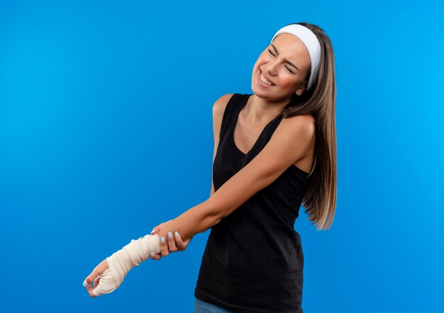 Douleur jeune fille assez sportive portant un bandeau et un bracelet tenant son poignet blessé enveloppé d'un bandage avec les yeux fermés isolé sur le mur bleu