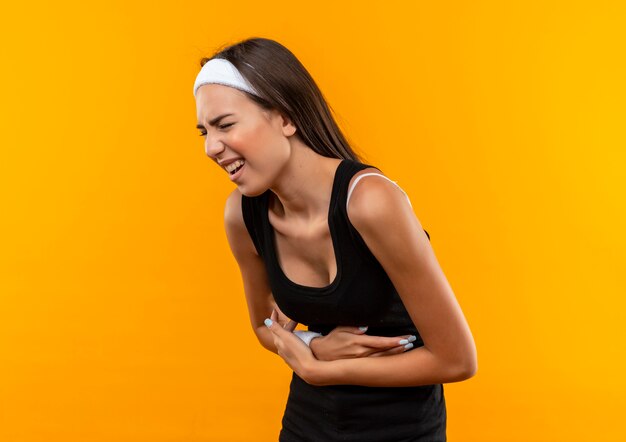 Douleur jeune fille assez sportive portant un bandeau et un bracelet tenant son ventre souffrant de douleur avec les yeux fermés isolés sur le mur orange