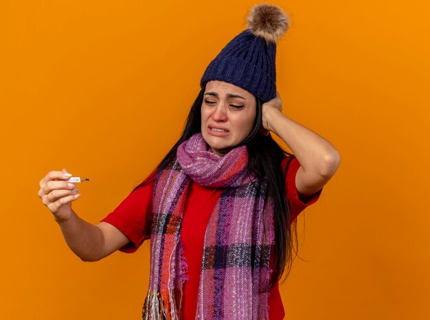 Douleur jeune fille malade caucasienne portant chapeau d'hiver et écharpe tenant et regardant thermomètre mettre la main sur la tête isolé sur un mur orange avec espace de copie