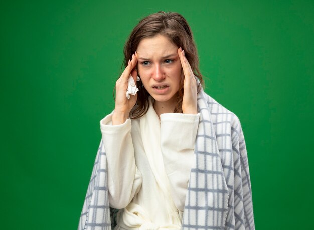 Douleur jeune fille malade à côté portant une robe blanche enveloppée de plaid mettant les mains sur les tempes isolées sur le vert