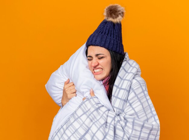 Douleur jeune fille malade de race blanche portant un chapeau d'hiver et une écharpe enveloppée dans un oreiller étreignant à carreaux avec les yeux fermés isolé sur fond orange avec espace de copie