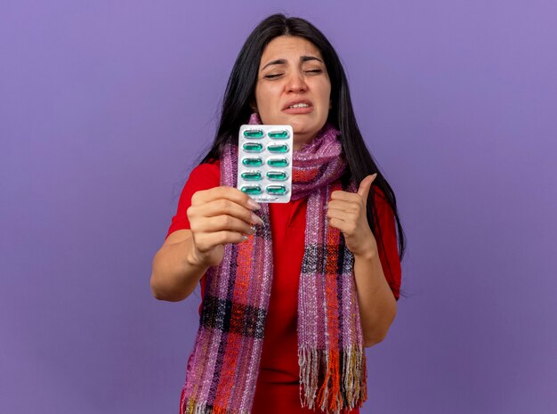 Douleur jeune fille malade de race blanche portant un foulard montrant pack de capsules et le pouce vers le haut avec les yeux fermés isolé sur mur violet avec espace copie