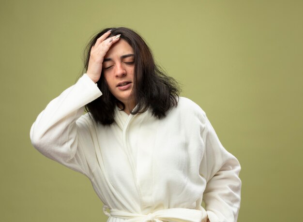 Douleur jeune fille malade de race blanche portant robe en gardant la main sur la tête avec les yeux fermés isolé sur fond vert olive