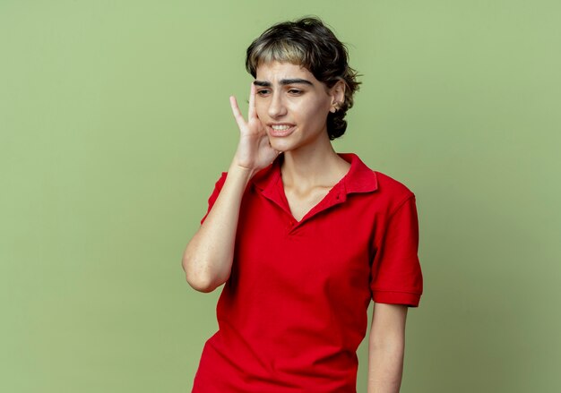 Douleur jeune fille de race blanche avec coupe de cheveux de lutin mettant la main sur le temple regardant vers le bas souffrant de maux de tête isolé sur fond vert olive avec espace de copie