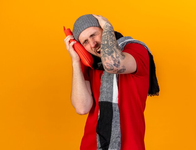 Douleur jeune homme malade de race blanche portant un chapeau d'hiver et une écharpe tient la tête et met une bouteille d'eau chaude isolée sur un mur orange avec espace de copie