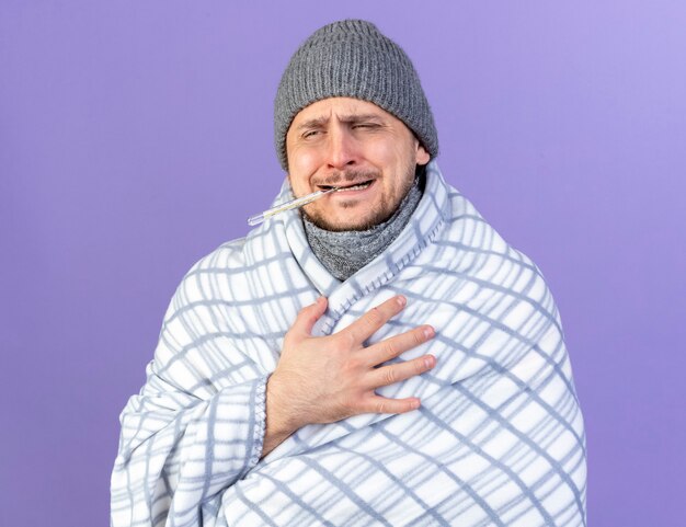 Douleur jeune homme slave malade blonde portant un chapeau d'hiver et une écharpe enveloppée dans un plaid détient un thermomètre avec des dents isolées sur un mur violet avec espace de copie