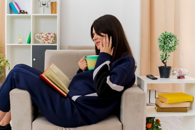 Douleur jeune jolie femme caucasienne assise sur un fauteuil dans un salon conçu tenant une tasse avec un livre sur les jambes mettant la main sur la joue souffrant de maux de dents livre de lecture