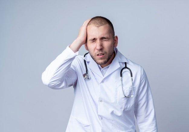 Douleur jeune médecin de sexe masculin portant une robe médicale et un stéthoscope autour de son cou mettant la main sur la tête avec les yeux fermés isolé sur blanc