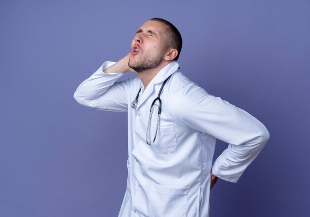 Douleur jeune médecin de sexe masculin portant une robe médicale et stéthoscope debout en vue de profil mettant les mains sur le cou et le dos avec les yeux fermés isolés sur violet avec espace copie