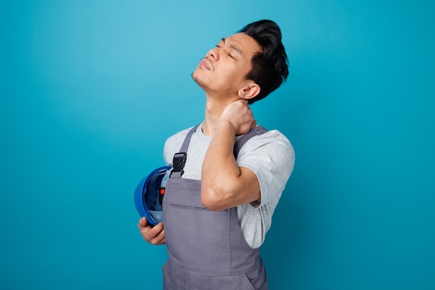 Douleur jeune travailleur de la construction portant un casque de sécurité et uniforme debout en vue de profil en gardant la main sur le cou tenant un casque de sécurité avec les yeux fermés