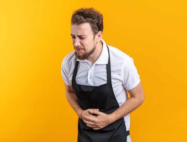 Douleur avec les yeux fermés jeune homme barbier vêtu d'un estomac attrapé uniforme isolé sur mur jaune