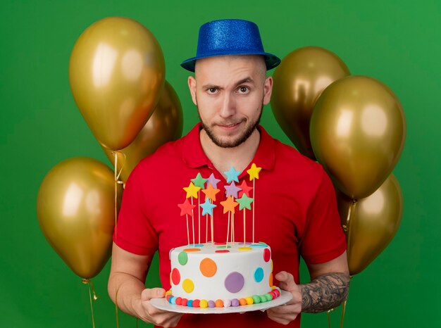 Doute jeune beau mec de fête portant chapeau de fête debout devant des ballons tenant le gâteau d'anniversaire à l'avant isolé sur mur vert