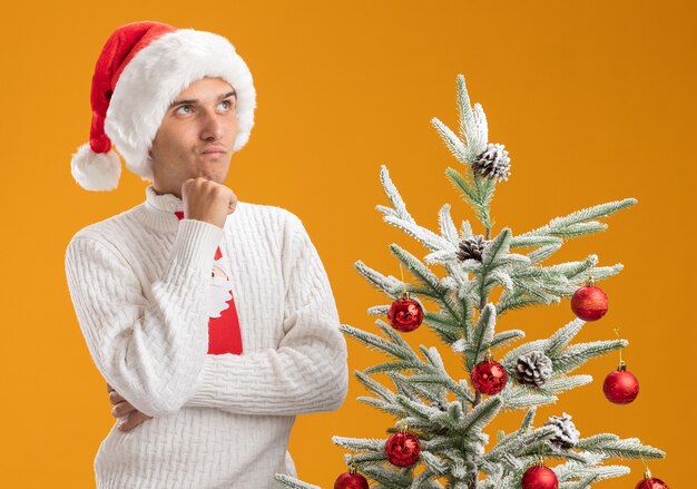 Doute jeune beau mec portant chapeau de Noël et cravate de père Noël debout avec une posture fermée près de sapin de Noël décoré en levant isolé sur fond orange
