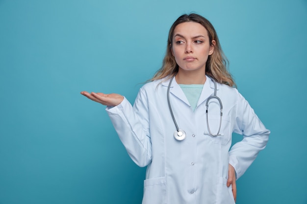 Doute jeune femme médecin portant une robe médicale et un stéthoscope autour du cou en gardant la main sur la taille montrant la main vide à côté