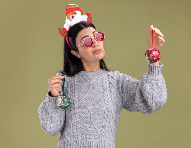 Photo gratuite doute jeune fille caucasienne portant bandeau de père noël avec des lunettes tenant des boules de noël en regardant l'un d'eux isolé sur fond vert olive