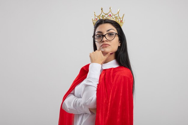 Doute jeune fille de super-héros caucasien portant des lunettes et une couronne debout en vue de profil en regardant la caméra toucher le menton isolé sur fond blanc avec espace copie