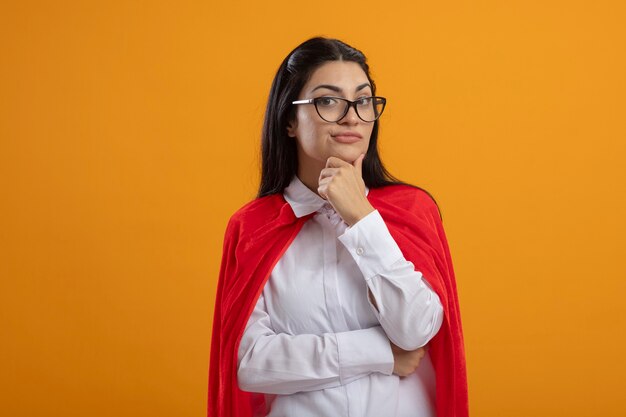 Doute jeune fille de super-héros caucasien portant des lunettes touchant le menton regardant la caméra isolée sur fond orange avec espace de copie