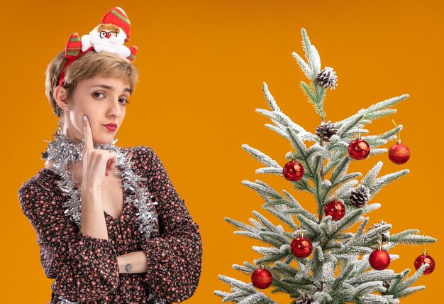Doute jeune jolie fille portant bandeau de père Noël et guirlande de guirlandes autour du cou debout près de l'arbre de Noël décoré touchant le visage isolé sur le mur orange