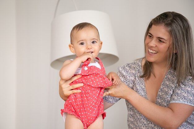 Doux bébé portant un corps rouge, debout avec le soutien des mamans, mordant la main et une partie des vêtements, souriant, a. Coup moyen. Concept de parentalité et d'enfance