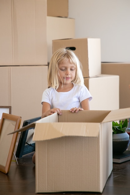 Doux cheveux blonds petite fille déballage des choses dans le nouvel appartement, assis sur le sol près de la boîte de dessin animé ouverte et regardant à l'intérieur