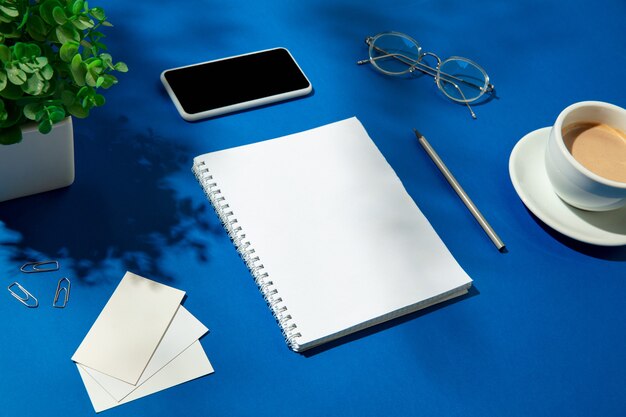 Draps, café et outils de travail sur une table bleue à l'intérieur. Lieu de travail créatif et confortable au bureau à domicile, maquette inspirante avec des ombres végétales à la surface. Concept de bureau distant, indépendant, ambiance.