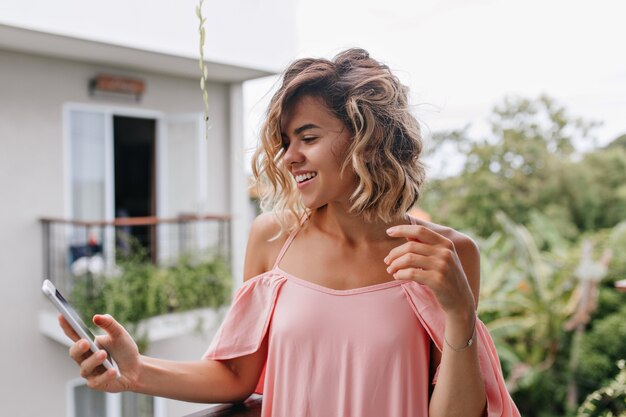 Dreamy fille bronzée texto message téléphonique avec le sourire. Incroyable modèle féminin bouclé debout au balcon de l'hôtel et regardant l'écran du smartphone.