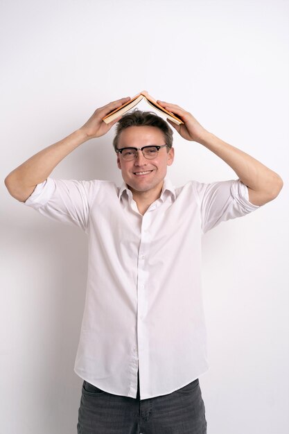 Drôle beau mec dans des verres se cachant sous son livre préféré le tenant au-dessus de la tête avec un charmant sourire positif sur son visage porter une chemise blanche et un jean gris isolé concept d'apprentissage sur fond blanc