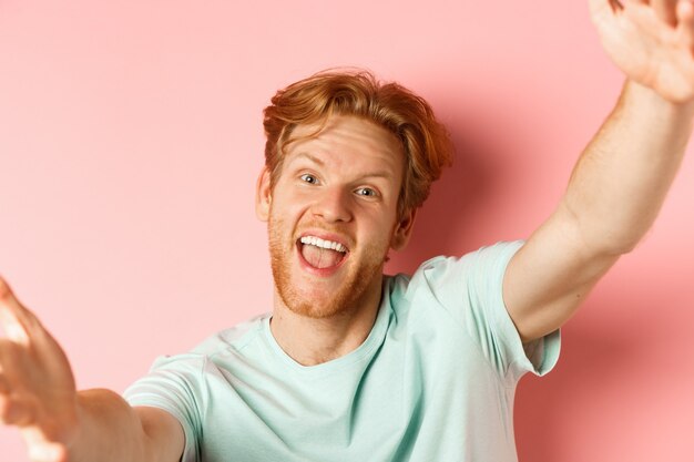 Drôle bel homme rousse prenant selfie, étendez les mains pour tenir la caméra et souriant heureux, regardez depuis un smartphone, debout sur fond rose