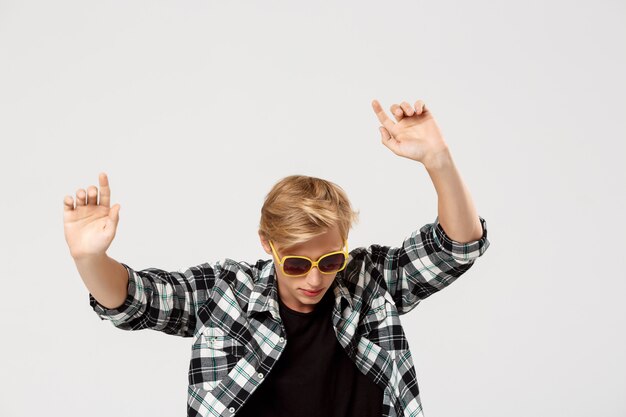 Drôle blond beau jeune homme portant des lunettes de soleil et chemise à carreaux décontractée dansant en agitant les mains l'air sur le mur gris