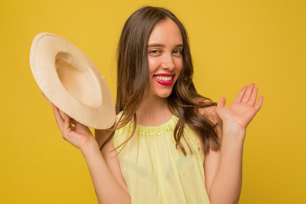 Drôle charmante fille aux lèvres roses renonce à la caméra tout en tenant un chapeau et en se présentant à la caméra sur fond jaune