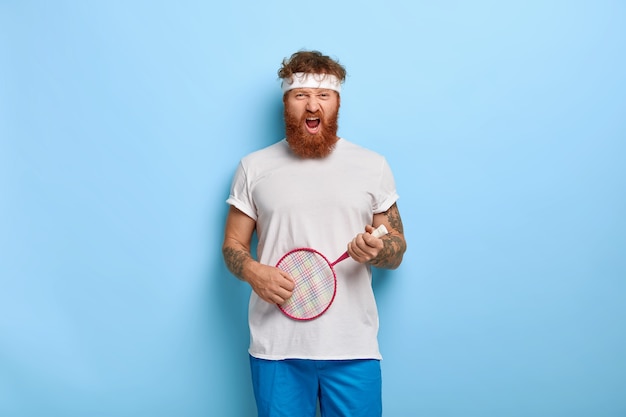 Drôle déterminé joueur de tennis aux cheveux rouges tient la raquette posant contre le mur bleu